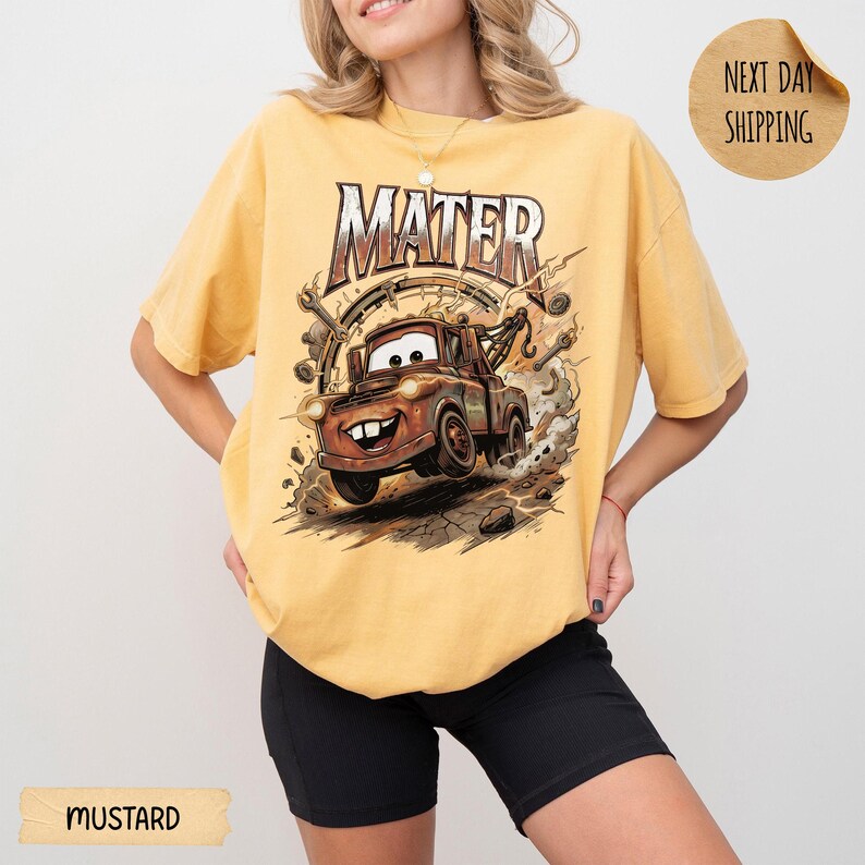 May include: Mustard-coloured t-shirt with a cartoon image of Mater from the Cars film. The graphic includes the name "Mater" in a stylized font, along with Mater's image, surrounded by tools and a cracked ground effect.
