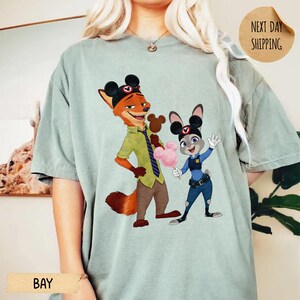 May include: A light green t-shirt featuring a cartoon illustration of Nick Wilde and Judy Hopps from Zootopia. Nick is wearing a green shirt and brown pants, while Judy is in a police uniform. The shirt has the word "BAY" printed on it.