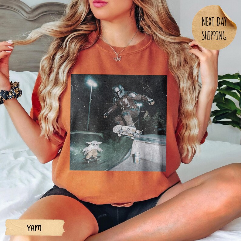 May include: Burnt orange t-shirt featuring a graphic of a character from a popular sci-fi series skateboarding, with a smaller character also on a skateboard. The image is set in a skate park at night. The shirt has the word "YAM" printed on it.