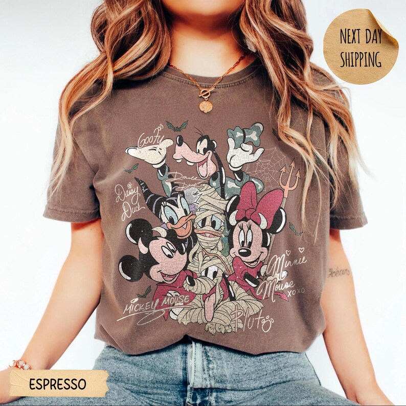 Peut inclure: T-shirt couleur expresso avec un motif de personnages Disney en costumes d'Halloween. Mickey Mouse, Minnie Mouse, Donald Duck, Daisy Duck, Dingo et Pluto sont repr&eacute;sent&eacute;s. Le t-shirt a un aspect vintage et vieilli.