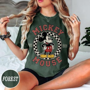 Puede incluir: Camiseta verde bosque con un dise&ntilde;o vintage de Mickey Mouse. El dise&ntilde;o incluye las palabras "Mickey Mouse" en rojo, un borde a cuadros y el a&ntilde;o "1928". La camiseta tiene cuello redondo y mangas cortas.
