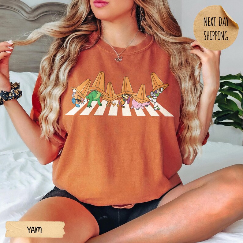 May include: A burnt orange crewneck sweatshirt featuring a graphic print of five cartoon characters walking in a line across a white striped crosswalk, each wearing an orange traffic cone on their head. The fabric is a solid, muted orange colour.