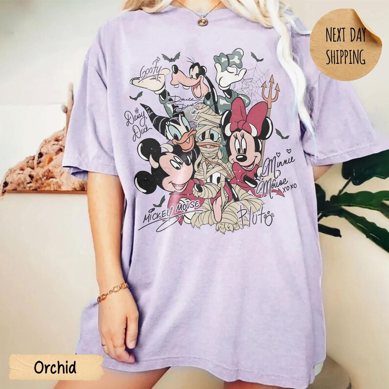 Peut inclure: T-shirt de couleur orchid&eacute;e avec un motif de personnages Disney, dont Mickey Mouse, Minnie Mouse et Dingo, en costumes d'Halloween. Le t-shirt porte l'inscription "Mickey Mouse" et "Minnie Mouse". Un autocollant rond indique "Next Day Shipping".