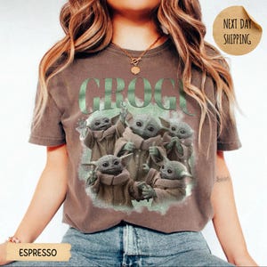 May include: A brown short-sleeve t-shirt featuring a graphic print of five small green creatures with large ears and eyes. The text GROGU is printed in large green letters above the creatures. The shirt has a crew neckline and a relaxed fit.