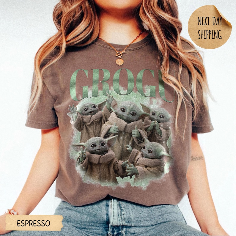 May include: A brown short-sleeve t-shirt featuring a graphic print of five small green creatures with large ears and eyes. The text GROGU is printed in large green letters above the creatures. The shirt has a crew neckline and a relaxed fit.