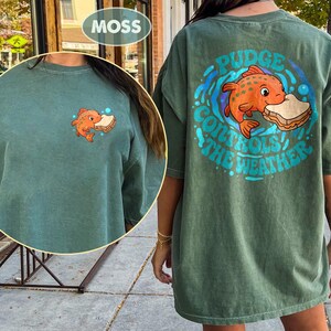 May include: A moss green t-shirt featuring a cartoon orange fish with a sandwich, and the text "Pudge Controls The Weather" in a blue circle on the back. A smaller fish graphic is on the front.
