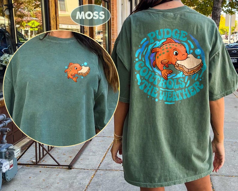 May include: A moss green t-shirt featuring a cartoon orange fish with a sandwich, and the text "Pudge Controls The Weather" in a blue circle on the back. A smaller fish graphic is on the front.