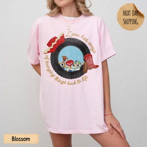 May include: A light pink t-shirt featuring a graphic of a black tire surrounding a blue sky scene with cartoon characters. A red cowboy hat and a red cowboy boot are positioned on the tire. The text reads, Love has ways of bringing things back to life.