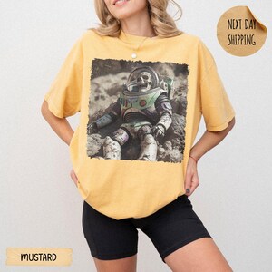 May include: A mustard yellow short-sleeve t-shirt featuring a graphic print of a space ranger action figure with a skull head sitting on a rocky, cratered surface. The design is centered on the chest of the garment, which has a crew neckline.
