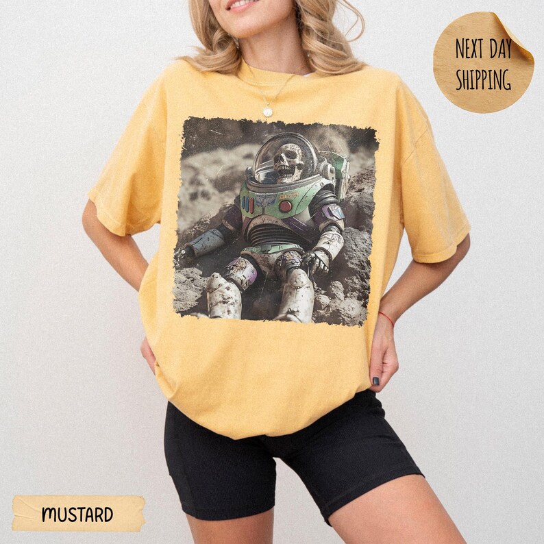 May include: A mustard yellow short-sleeve t-shirt featuring a graphic print of a space ranger action figure with a skull head sitting on a rocky, cratered surface. The design is centered on the chest of the garment, which has a crew neckline.