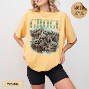 May include: A mustard yellow short-sleeve t-shirt featuring a graphic print of five Grogu characters in various poses. The text GROGU is printed in large green letters above the characters. The shirt has a crew neckline and a relaxed fit.