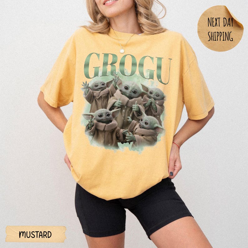 May include: A mustard yellow short-sleeve t-shirt featuring a graphic print of five Grogu characters in various poses. The text GROGU is printed in large green letters above the characters. The shirt has a crew neckline and a relaxed fit.