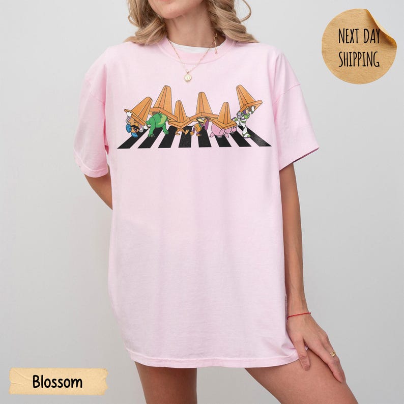May include: A light pink t-shirt featuring a graphic of five cartoon characters walking across a black and white striped crosswalk. Each character has an orange traffic cone placed on their head. The design is centred on the chest area.