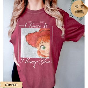 May include: A crimson short-sleeve t-shirt featuring a graphic print of a cartoon character with red hair and a red hat inside a square frame. The text reads "EST 2026", "I Knew It", and "I Knew You" with a small musical treble clef symbol below.