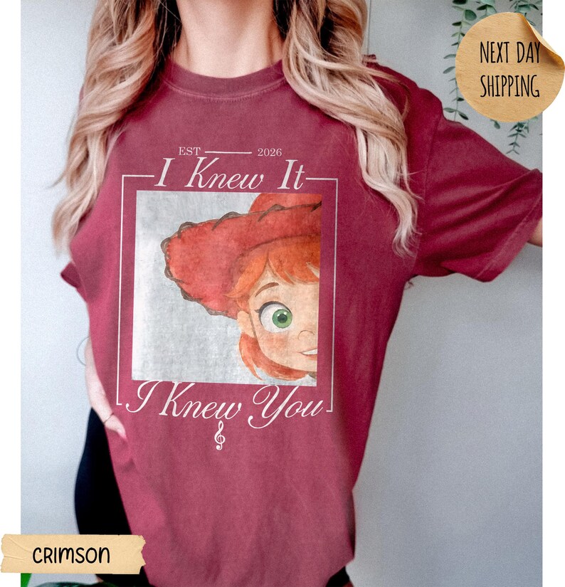 May include: A crimson short-sleeve t-shirt featuring a graphic print of a cartoon character with red hair and a red hat inside a square frame. The text reads "EST 2026", "I Knew It", and "I Knew You" with a small musical treble clef symbol below.