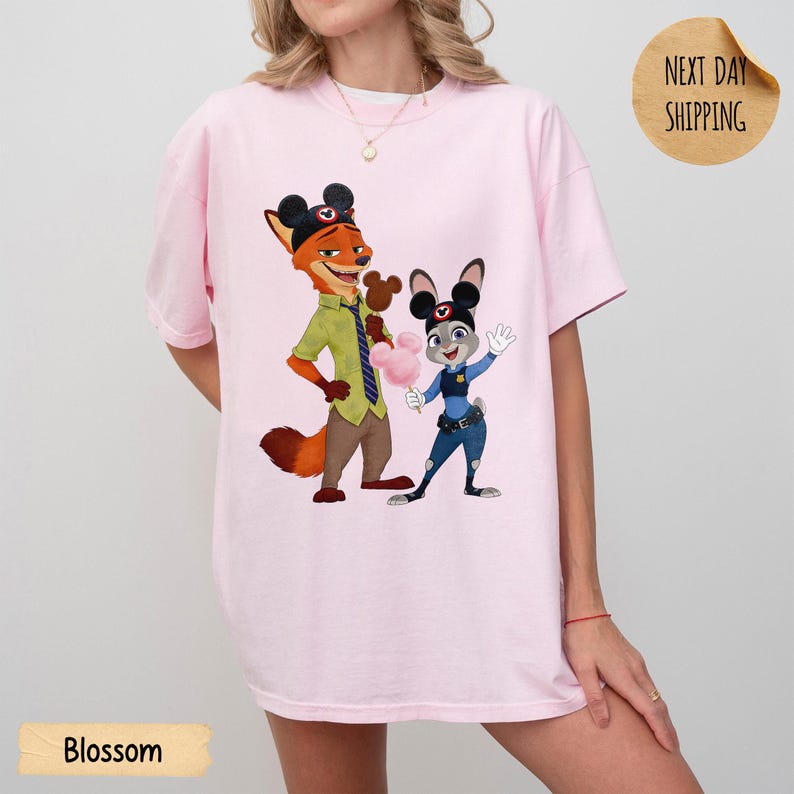 May include: A light pink t-shirt featuring a cartoon graphic of Nick Wilde and Judy Hopps from Zootopia. Nick is wearing a green shirt and Judy is in a police uniform. The word "Blossom" is printed at the bottom of the shirt.