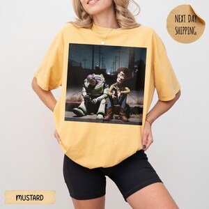 Pode incluir: Camiseta mostarda com um gr&aacute;fico de Buzz Lightyear e Woody de Toy Story sentados em uma cal&ccedil;ada. A imagem tem um fundo urbano escuro. A camiseta tem um caimento folgado.