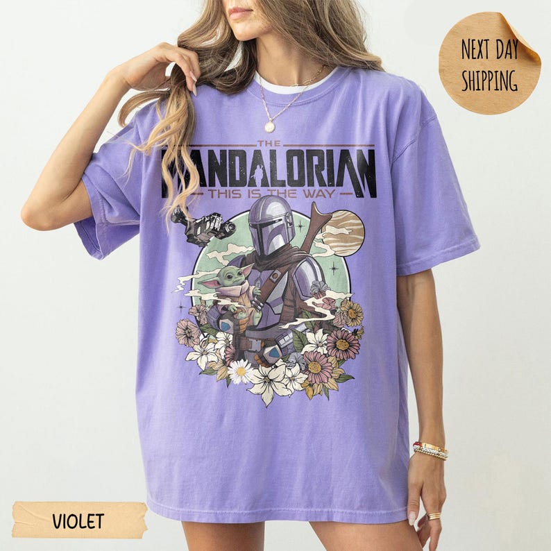 May include: Violet-colored t-shirt featuring a graphic of The Mandalorian and Grogu surrounded by flowers. The text on the shirt reads "THE MANDALORIAN THIS IS THE WAY". The shirt has short sleeves and a crew neck.