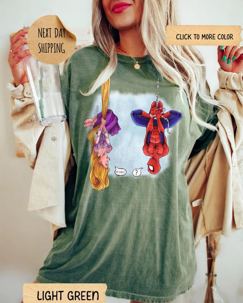 Op de afbeelding: Een lichtgroen t-shirt met een grafische print van een blond personage met lang haar dat ondersteboven hangt en een rood-blauwe superheld die ondersteboven aan een ketting hangt. De tekst O HAI en een vraagteken staan tussen de personages.