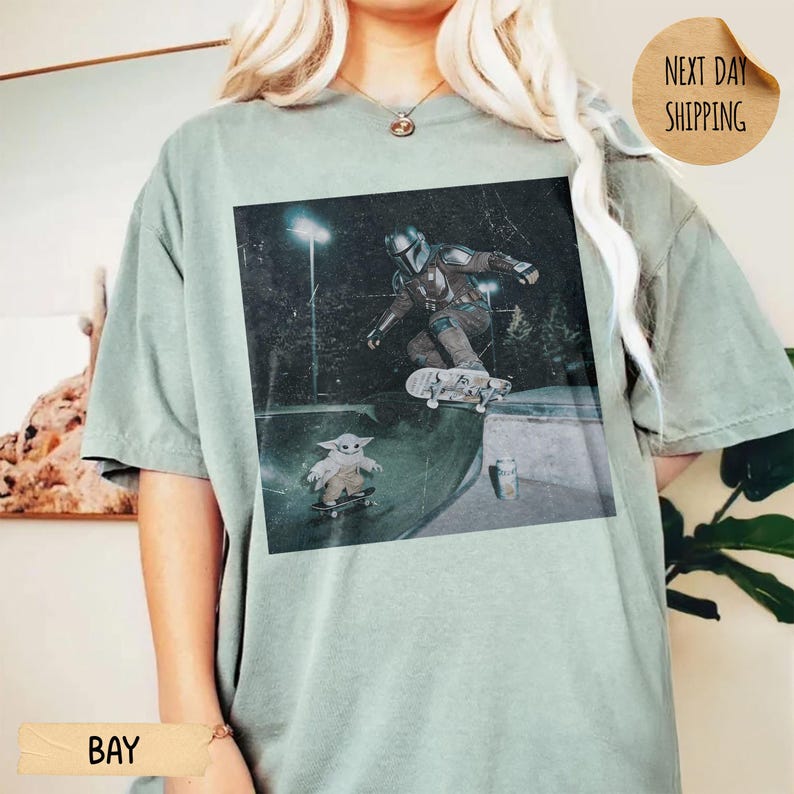 May include: Sage green t-shirt featuring a graphic of The Mandalorian and Grogu skateboarding in a skate park. The image has a vintage, faded look. The shirt also has the text "BAY" and "NEXT DAY SHIPPING".