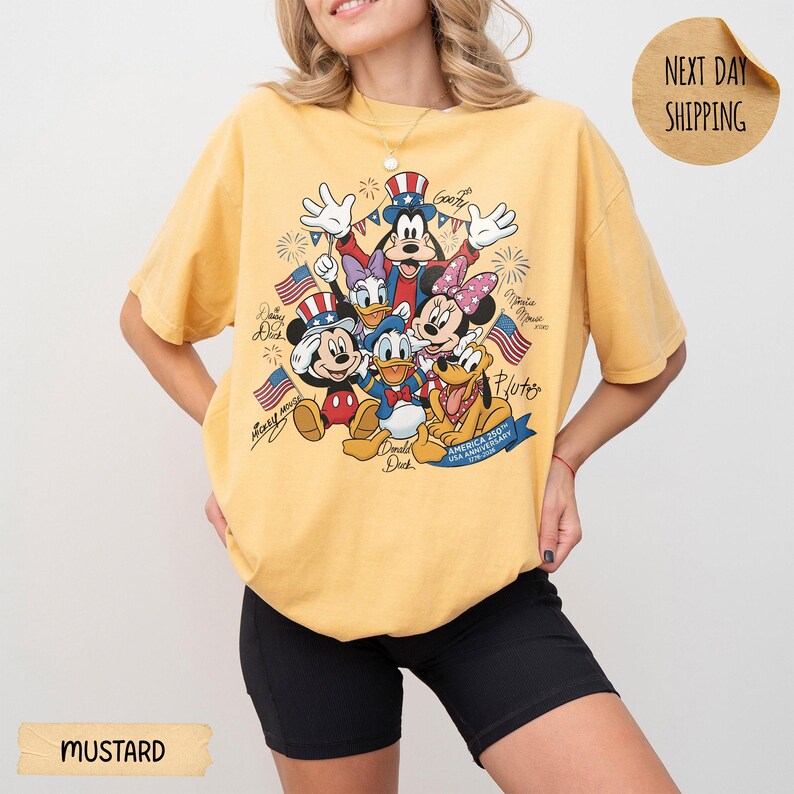May include: A mustard yellow t-shirt featuring a graphic print of Disney characters including Mickey Mouse, Minnie Mouse, Donald Duck, Daisy Duck, Goofy, and Pluto wearing patriotic hats. The text reads America 250th USA Anniversary 1776-2026.