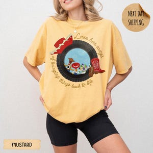 May include: A mustard yellow short-sleeve t-shirt featuring a graphic of a black tire framing cartoon characters, a red cowboy hat, and a red cowboy boot. The text reads, Love has ways of bringing things back to life, in a circular script font.