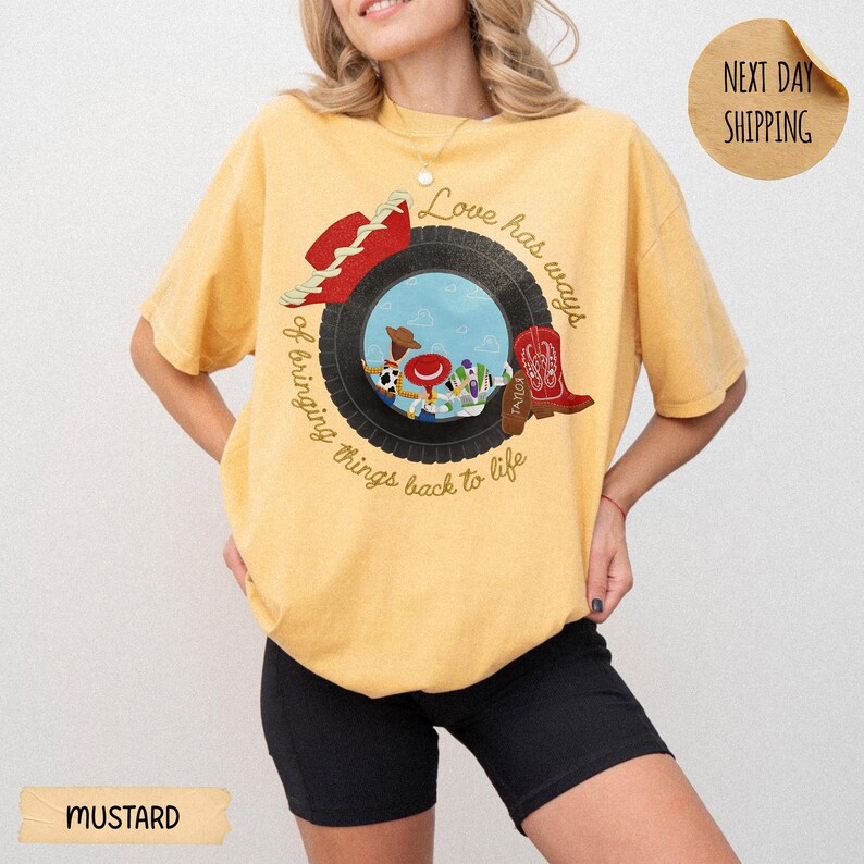 May include: A mustard yellow short-sleeve t-shirt featuring a graphic of a black tire framing cartoon characters, a red cowboy hat, and a red cowboy boot. The text reads, Love has ways of bringing things back to life, in a circular script font.