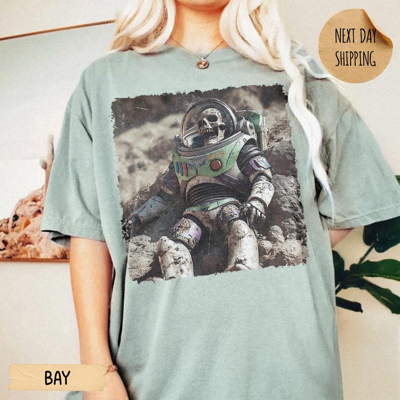 May include: A sage green short-sleeve t-shirt featuring a graphic print of a skeleton wearing a space suit with a clear helmet. The astronaut figure is positioned on a rocky, textured surface. The shirt has a crew neckline and a relaxed fit.