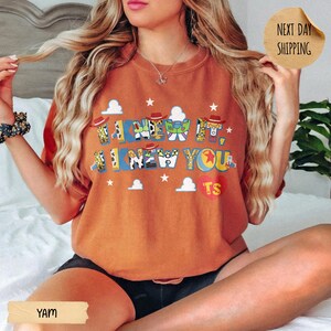 May include: A burnt orange crewneck t-shirt featuring a graphic print with the text I KNEW IT, I KNEW YOU. The letters are filled with patterns inspired by toy characters, including cow print, space ranger colors, and a red heart with the letters TS.