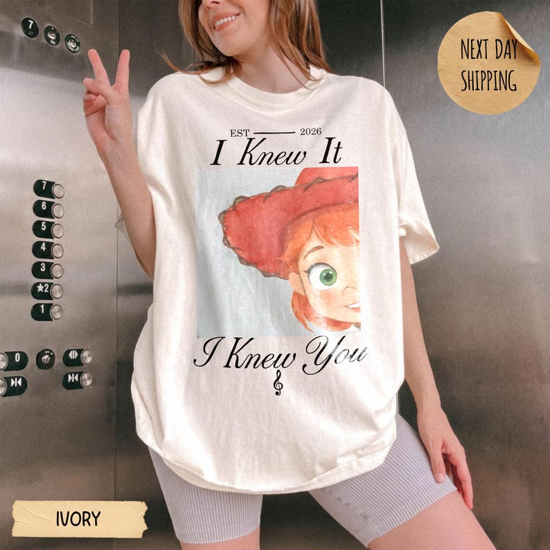 May include: An ivory t-shirt featuring a graphic of a cartoon character with red hair and a red hat. The text reads "EST 2026 I Knew It I Knew You" with a small treble clef symbol below the text. The design is centered on the front of the shirt.
