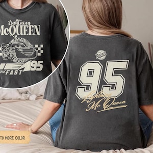May include: Dark gray t-shirt featuring Lightning McQueen graphics. The front displays the text "Lightning McQueen" above a car illustration and "95". The back shows a large "95" and "Lightning McQueen" in a vintage style.