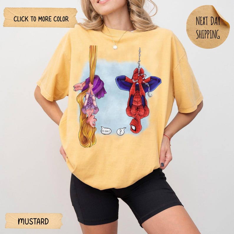 Op de afbeelding: Een mosterdgeel t-shirt met een grafische print van een langharig personage in een paarse jurk en een rood-blauw superheldenpersonage die beide ondersteboven hangen. Het personage in de paarse jurk zegt O HAI! en de superheld zegt ?.