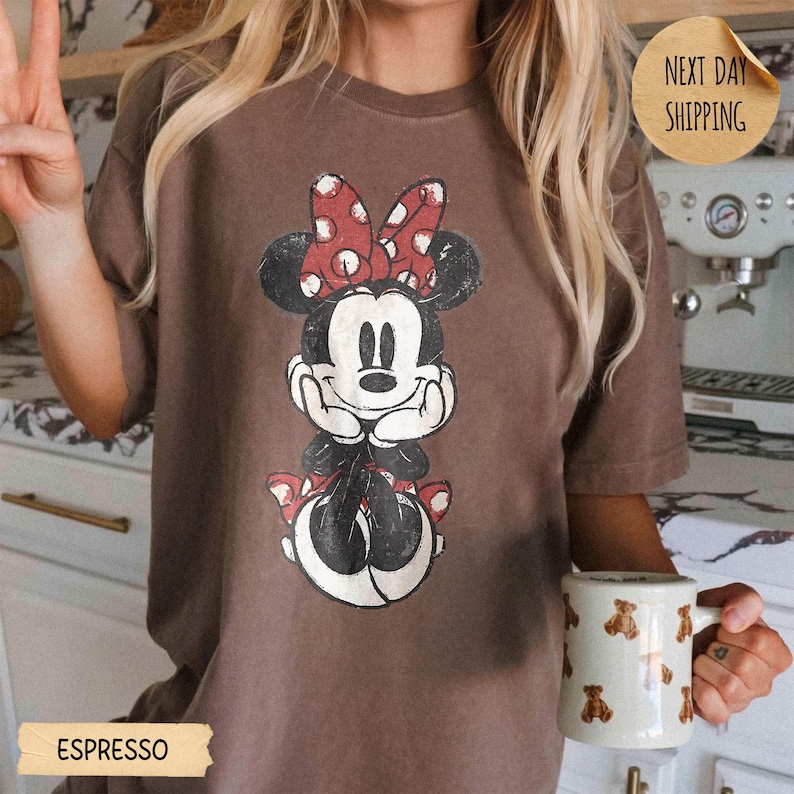 May include: A brown short-sleeve t-shirt featuring a distressed graphic of Minnie Mouse wearing a red and white polka dot bow. The shirt is paired with a white mug decorated with a repeating pattern of small brown teddy bears.