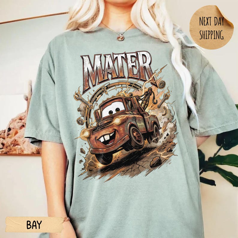 May include: A pale green t-shirt with a cartoon image of Mater from the film Cars. The word "MATER" is printed above the image in a brown, distressed font. The design includes a detailed illustration of Mater with a smiling face.