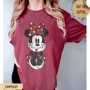 May include: A crimson short-sleeved t-shirt featuring a distressed graphic print of Minnie Mouse. The character is depicted with a red and white polka dot bow, black ears, and hands resting under the chin. The design has a vintage, worn-in aesthetic.