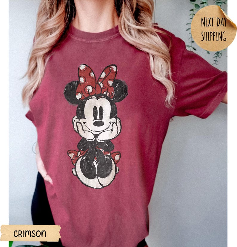May include: A crimson short-sleeved t-shirt featuring a distressed graphic print of Minnie Mouse. The character is depicted with a red and white polka dot bow, black ears, and hands resting under the chin. The design has a vintage, worn-in aesthetic.