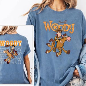 May include: Blue t-shirt featuring a cartoon illustration of Woody from Toy Story riding a horse, with the text "Sheriff Woody" above. The back of the shirt has the word "Woody" repeated multiple times with the same illustration.