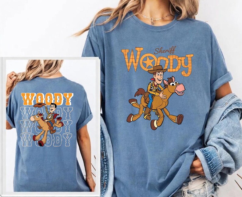 May include: Blue t-shirt featuring a cartoon illustration of Woody from Toy Story riding a horse, with the text "Sheriff Woody" above. The back of the shirt has the word "Woody" repeated multiple times with the same illustration.