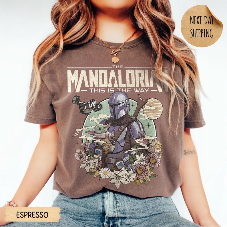 May include: Espresso-colored t-shirt featuring a graphic of The Mandalorian and Grogu surrounded by flowers. The text "THE MANDALORIAN - THIS IS THE WAY" is printed above the characters. The shirt has short sleeves.