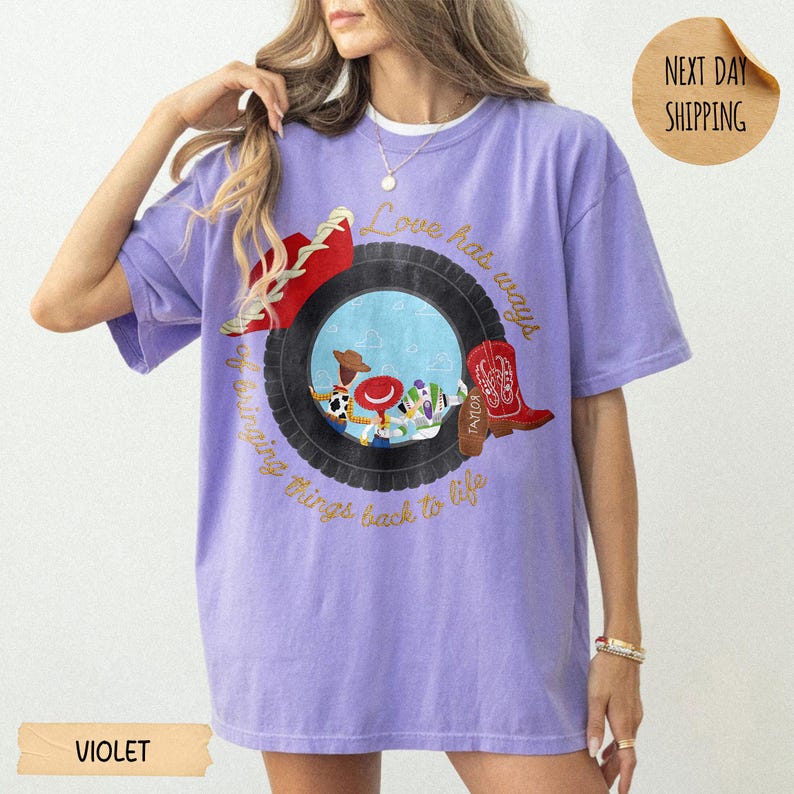 May include: A light purple short-sleeve t-shirt featuring a graphic of a black tire framing cartoon characters. The text reads Love has ways of bringing things back to life. A red cowboy boot and a red hat are positioned on the tire graphic.