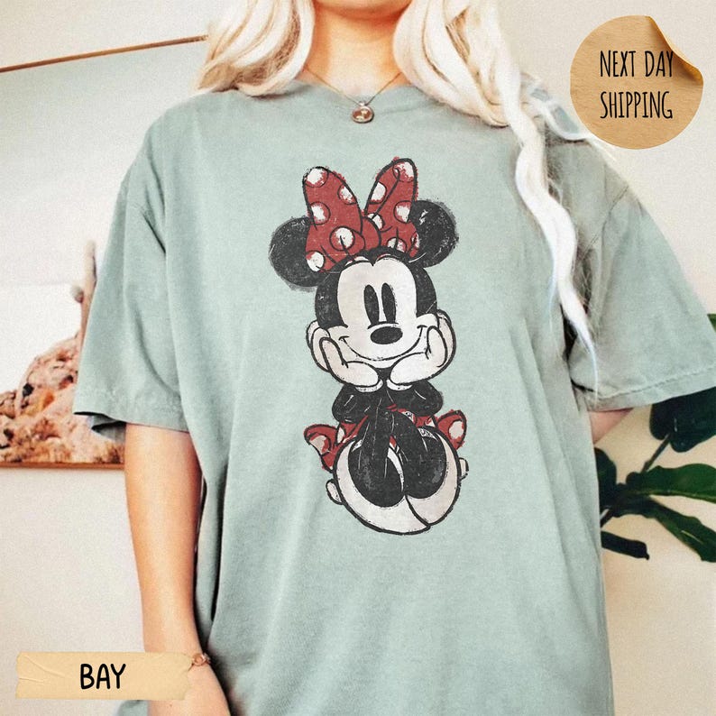 May include: A sage green short-sleeve t-shirt featuring a distressed graphic print of Minnie Mouse with her hands on her cheeks, wearing a red and white polka dot bow. The shirt has a crew neckline and a relaxed fit, showcasing a classic character design.