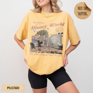 May include: Mustard yellow t-shirt featuring a vintage-style graphic of Disney World in Florida. The design includes Mickey Mouse, the Tree of Life, and Spaceship Earth, with the text "Florida Disney World" and "The Most Magical Place On Earth."