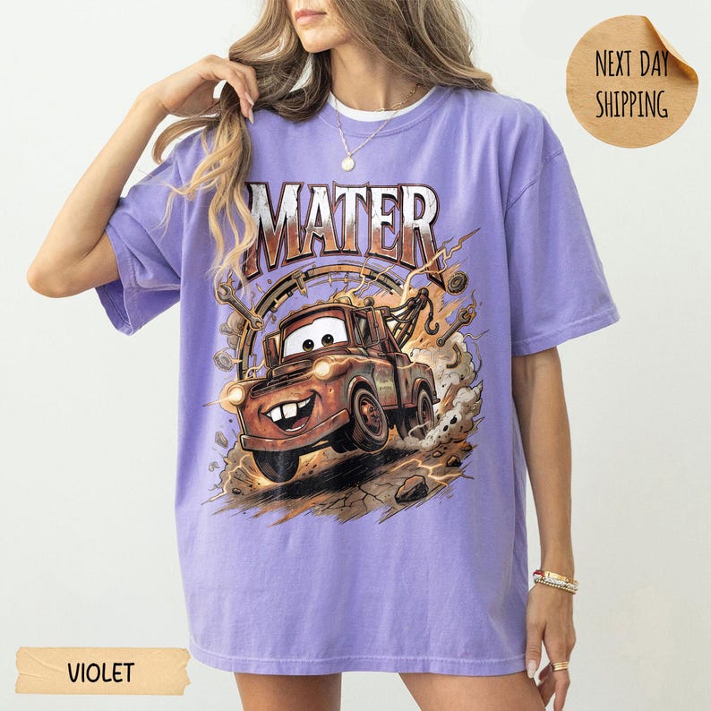 May include: Violet t-shirt with a graphic of Mater from the Cars film. The design features the word "MATER" above an illustration of the character. The shirt has short sleeves and a relaxed fit. The shirt is a shade of purple.