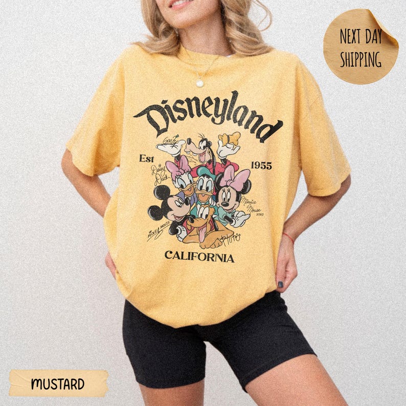 May include: A mustard yellow short-sleeve t-shirt featuring a graphic print of Disney characters including Mickey Mouse, Minnie Mouse, Donald Duck, Daisy Duck, and Goofy. The text reads Disneyland, Est 1955, and California in black lettering.