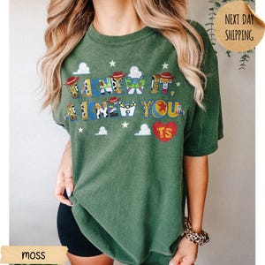 May include: A moss green short-sleeve t-shirt featuring a graphic print with the text I KNEW IT, I KNEW YOU. The letters are filled with patterns of cowboy hats, space ranger suits, and stars. A red heart with the letters TS is also printed.