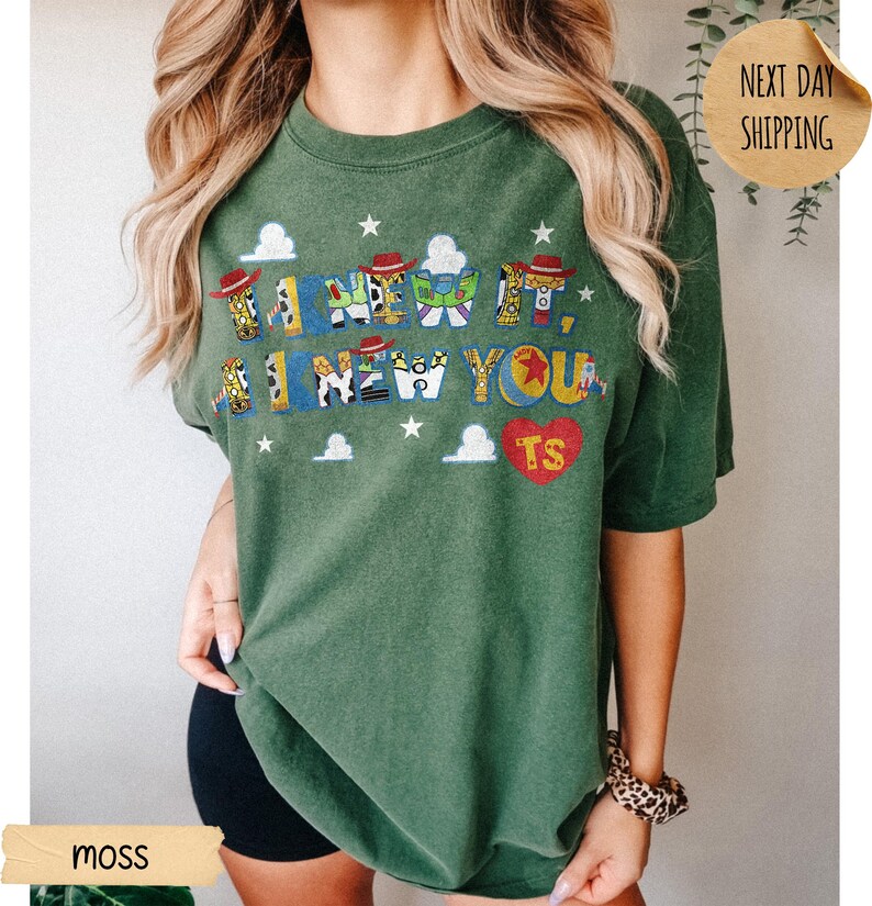 May include: A moss green short-sleeve t-shirt featuring a graphic print with the text I KNEW IT, I KNEW YOU. The letters are filled with patterns of cowboy hats, space ranger suits, and stars. A red heart with the letters TS is also printed.