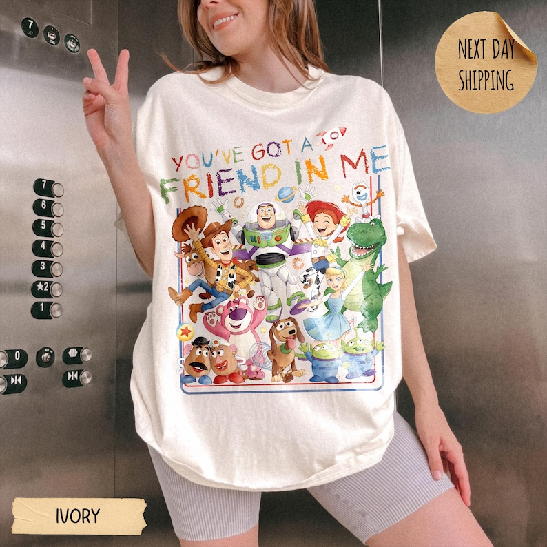 May include: An ivory t-shirt featuring a colorful graphic print of various animated characters from the Toy Story franchise. The text "YOU'VE GOT A FRIEND IN ME" is printed in multi-colored letters above the characters. The shirt has a crew neckline.
