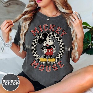 Puede incluir: Camiseta gris oscuro con un gr&aacute;fico vintage de Mickey Mouse. El dise&ntilde;o incluye las palabras "Mickey Mouse" en rojo, con un c&iacute;rculo a cuadros alrededor de Mickey. La camiseta tambi&eacute;n tiene el texto "EST. 1928".