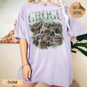 May include: A light purple short-sleeve t-shirt featuring a graphic print of five small green creatures in tan robes. The text GROGU is printed in large green letters above the creatures. The shirt has a crew neckline and a relaxed fit.