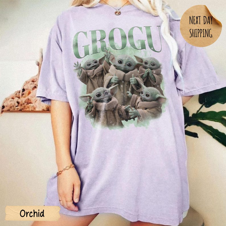May include: A light purple short-sleeve t-shirt featuring a graphic print of five small green creatures in tan robes. The text GROGU is printed in large green letters above the creatures. The shirt has a crew neckline and a relaxed fit.